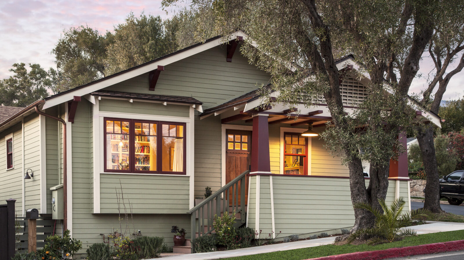 Exterior of a Craftsman style house with sage green siding and burgundy trim. Warm interior light glows through the picture window.