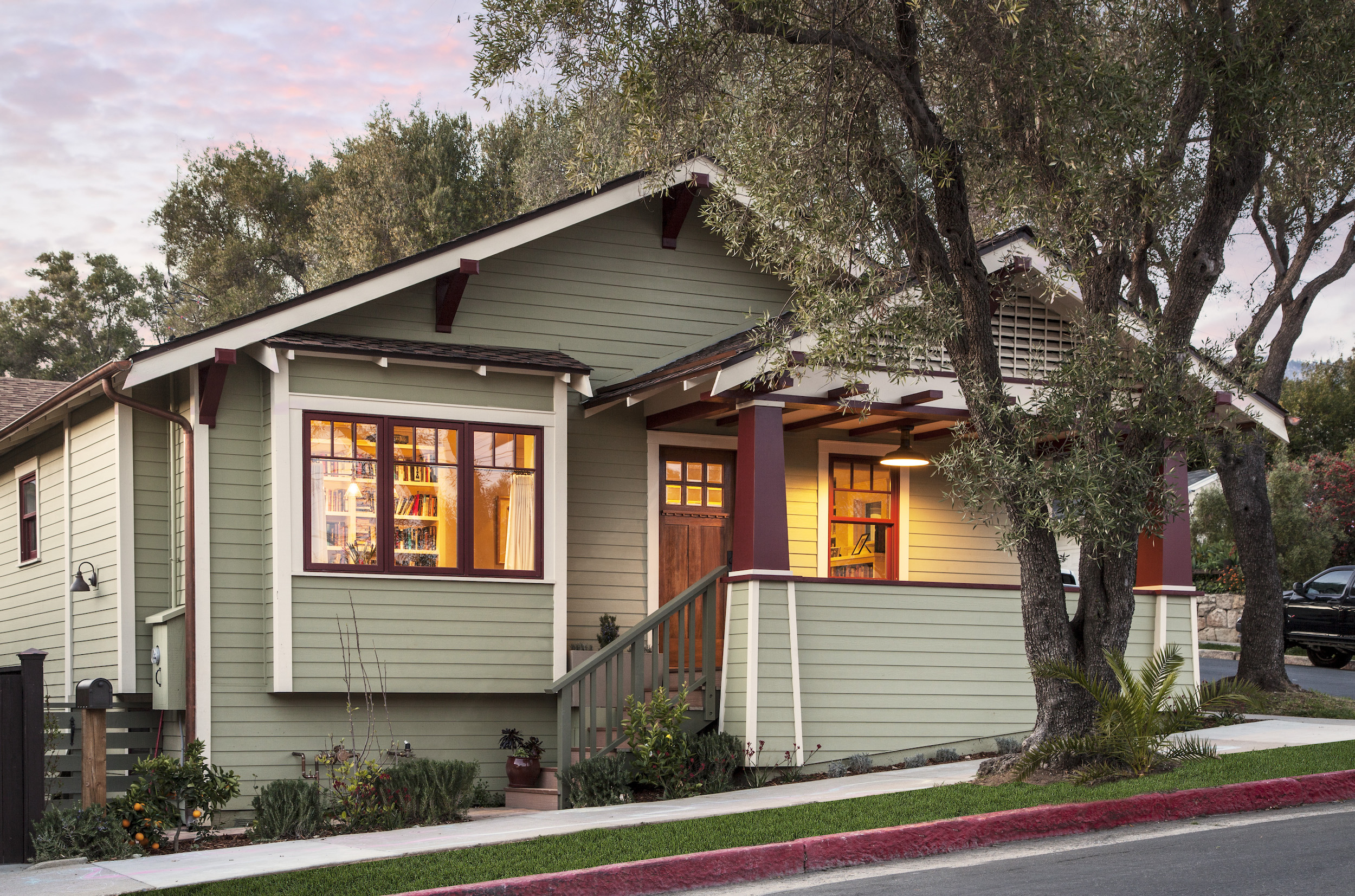 Exterior of a Craftsman style house with sage green siding and burgundy trim. Warm interior light glows through the picture window.