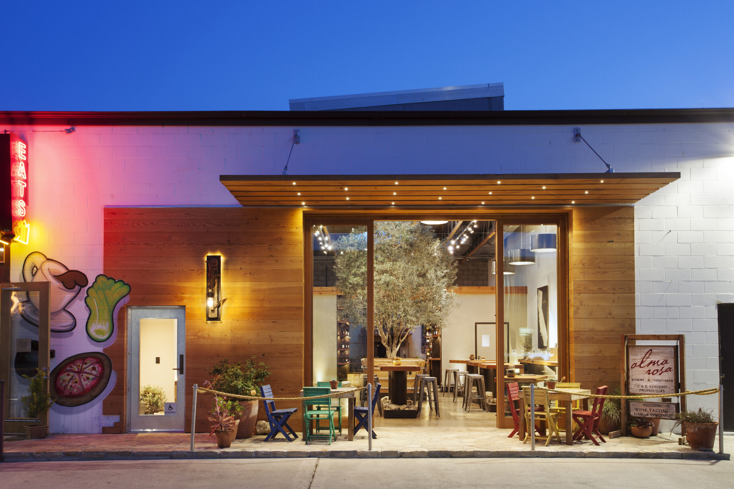 Exterior view of a modern tasting room at twilight, featuring large glass doors, a wooden awning, and colorful patio seating.