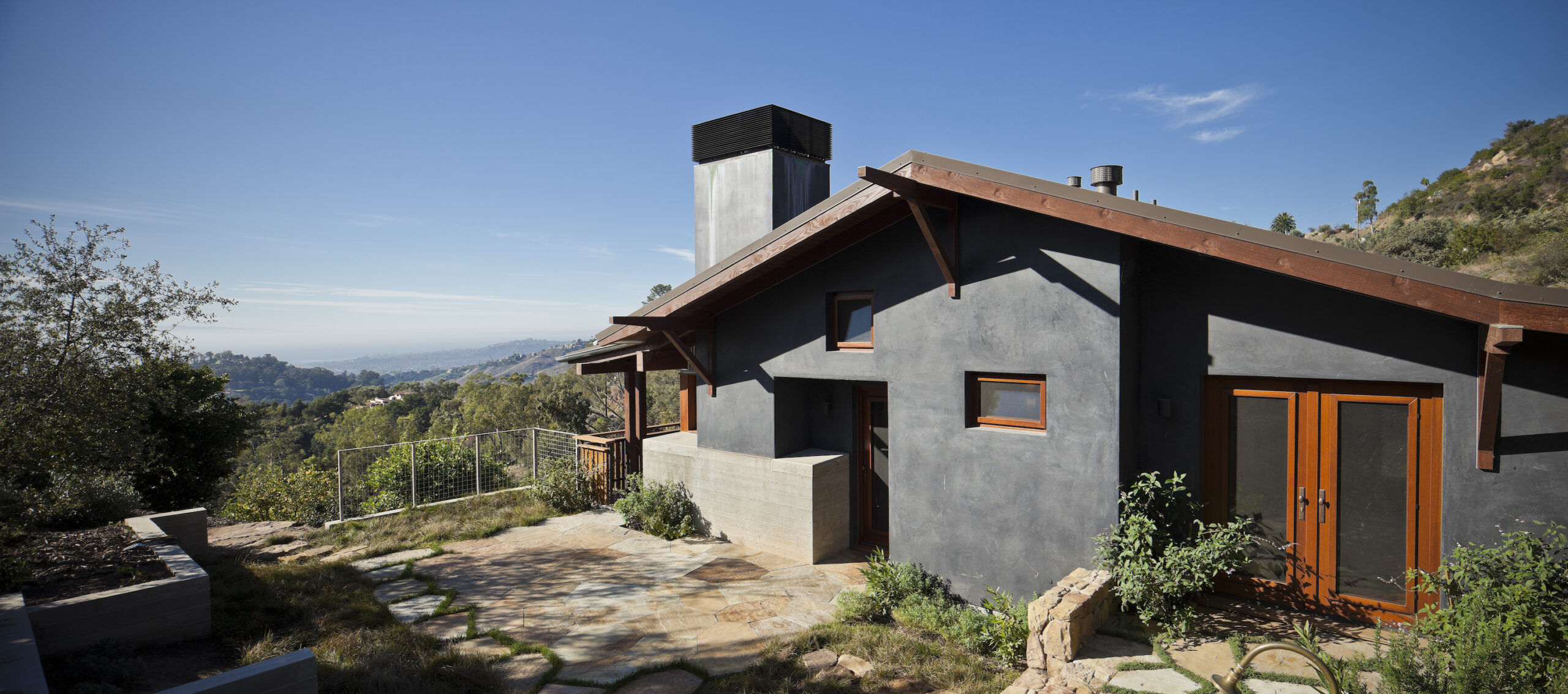 Modern hillside residence with dark stucco exterior, wood-framed windows and doors, and stone patio overlooking coastal views in Santa Barbara