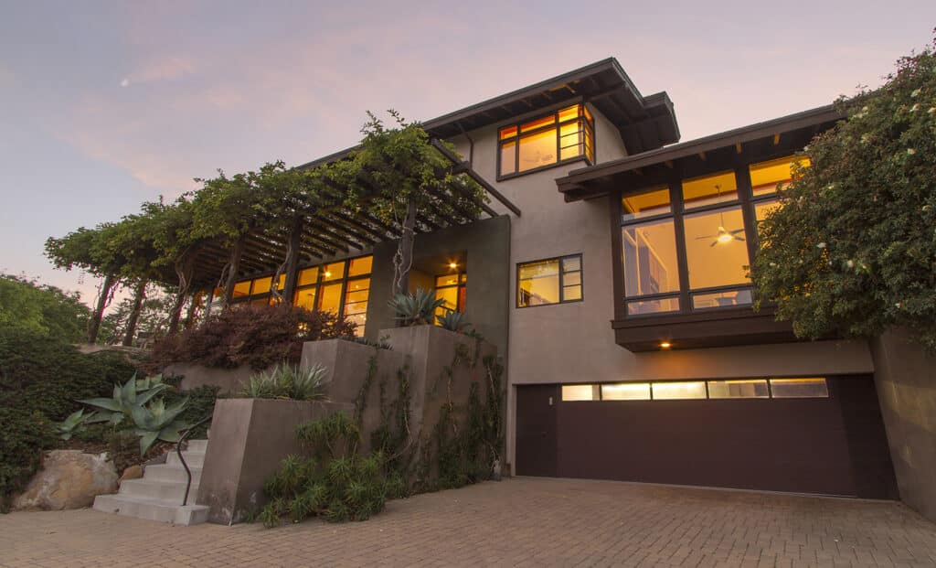Modern two-story home at dusk with warm interior lighting, large windows, a stone driveway, and lush landscaping.