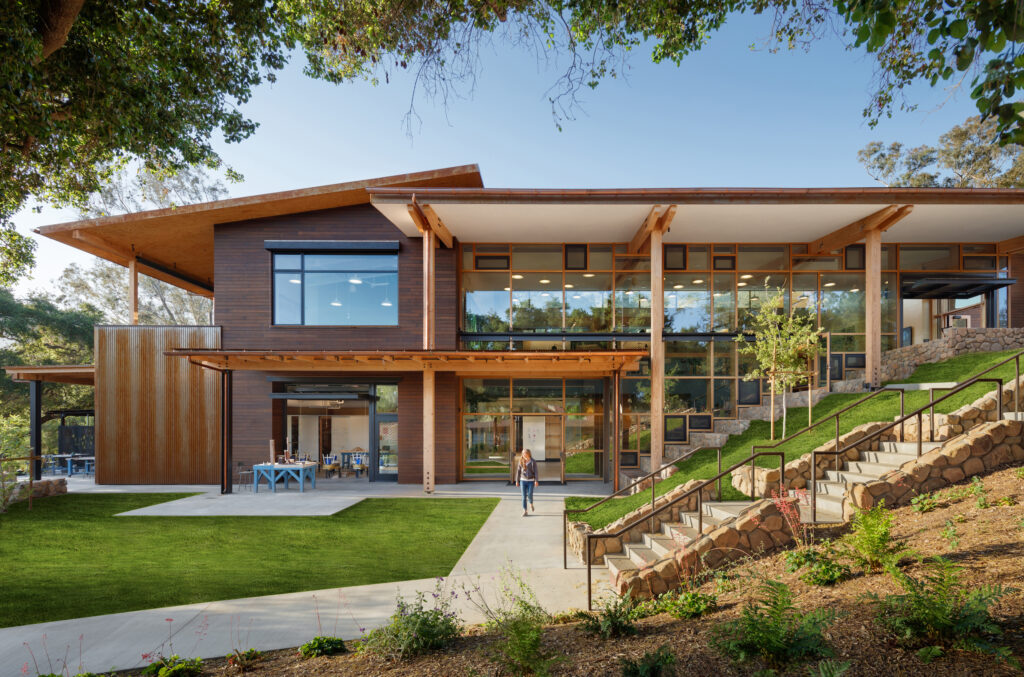 Exterior view of the Project-Based Learning Hub at The Thacher School. The design features a "transparent core" with floor-to-ceiling glass, exposed heavy timber columns, and a multi-level layout integrated into the natural Ojai topography with stone retaining walls and native landscaping.