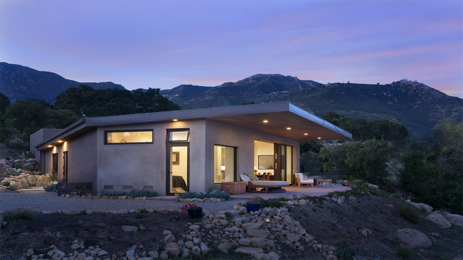 Modern single-story house with a flat roof and large windows, softly lit at dusk. The home sits on a rocky hillside surrounded by trees, with a covered patio and outdoor seating, and mountains rising in the background under a purple-blue evening sky