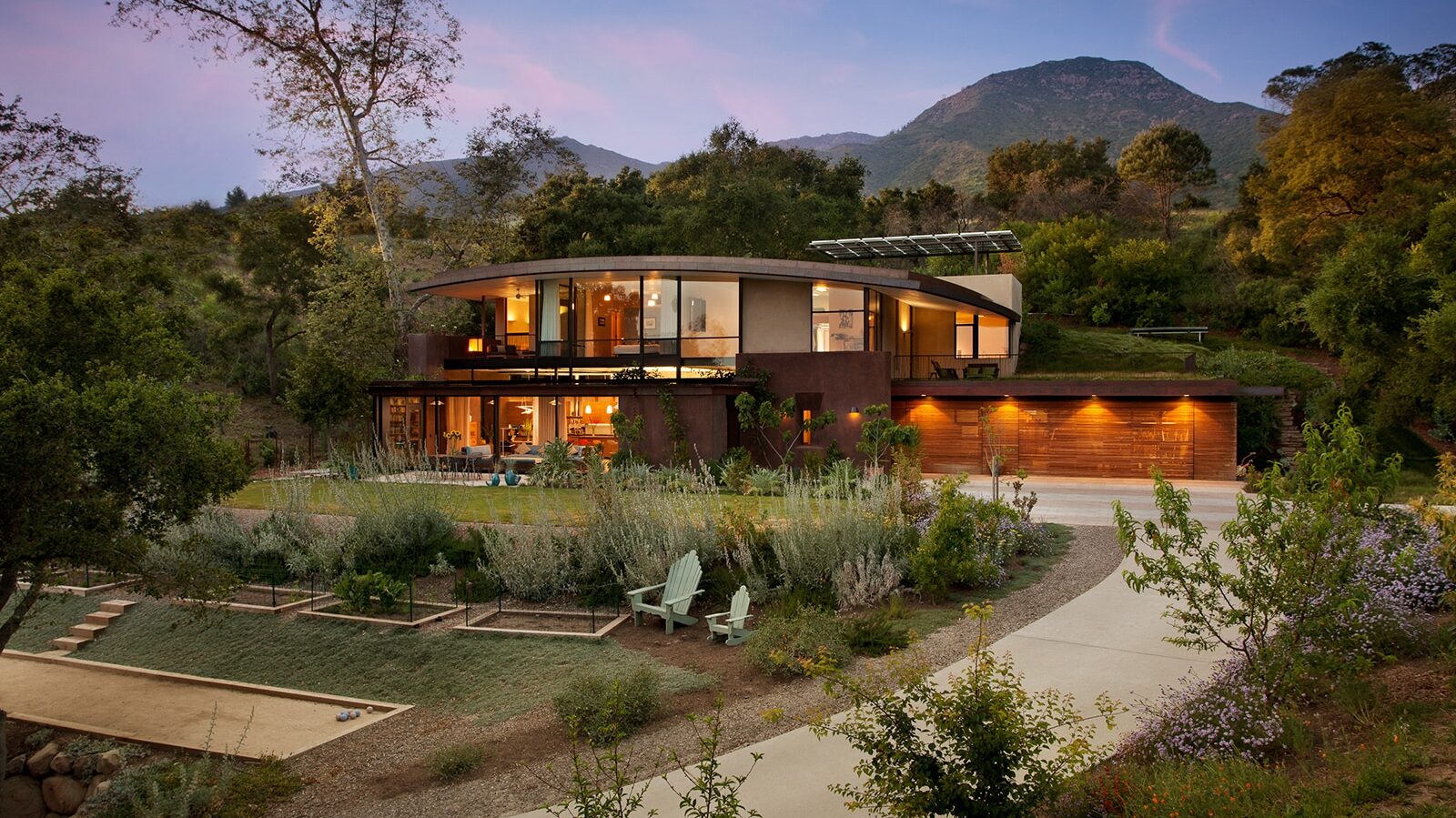 Contemporary hillside home with curved roof, large windows, and warm interior lighting.