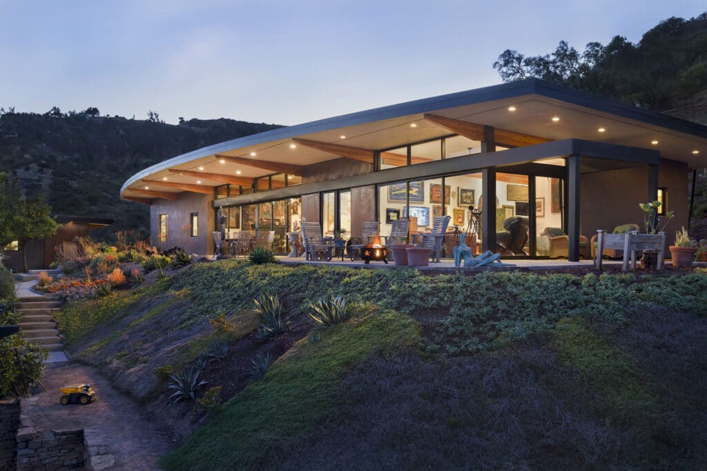 Dusk view of a modern hilltop home with a glowing interior, expansive glass walls, and a native plant garden.