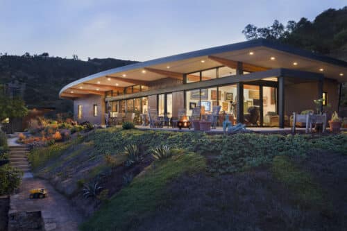 Dusk view of a modern hilltop home with a glowing interior, expansive glass walls, and a native plant garden.