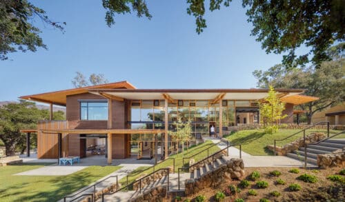 Exterior of the Thacher School Learning Hub, showcasing the sustainable "Western-vernacular" design. The photo highlights the transparent glass core, exposed heavy-timber framing, and the seamless transition between indoor maker spaces and the landscaped outdoor hillside.