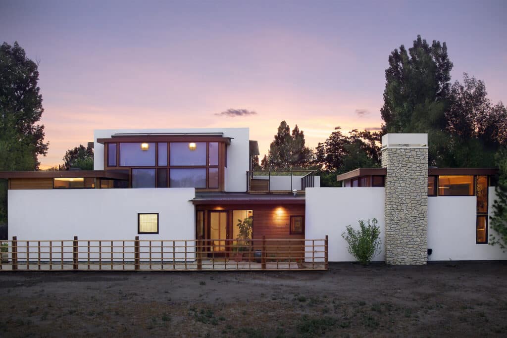 Wide view of a modern residence featuring a large stone chimney, white exterior walls, and expansive windows under a purple and orange sunset.