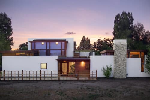 Wide view of a modern residence featuring a large stone chimney, white exterior walls, and expansive windows under a purple and orange sunset.