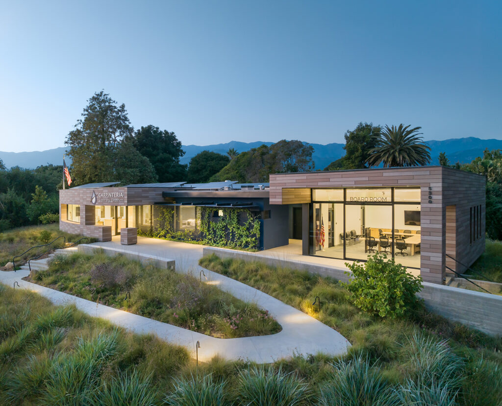 Modern administrative building nestled in native grassland. An elevated view of the Carpinteria Sanitary District building. It features a flat roof with solar panels, and a winding concrete path through drought-tolerant meadow grass.