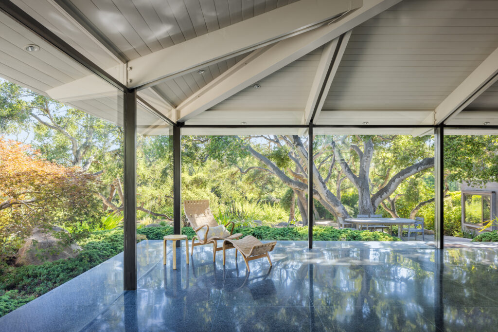 Midcentury open concept living room area with exposed structural beams, polished concrete floor, and lounge chair overlooking a landscaped garden