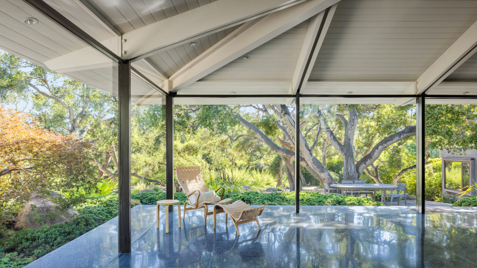 Midcentury open concept living room area with exposed structural beams, polished concrete floor, and lounge chair overlooking a landscaped garden