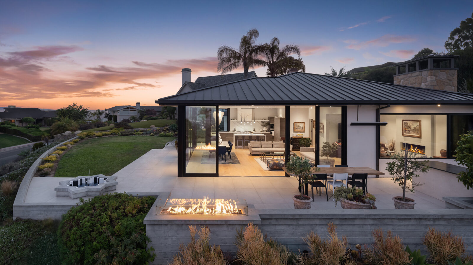 A photo of a house at dusk with the sunset in the background. The house has floor to ceiling windows. One of the windows is open and you can see inside the living room and dining area. In the forefront there is a terrace with an outdoor fire pit that has flames. There is a small water fountain on the left side of the terrace.