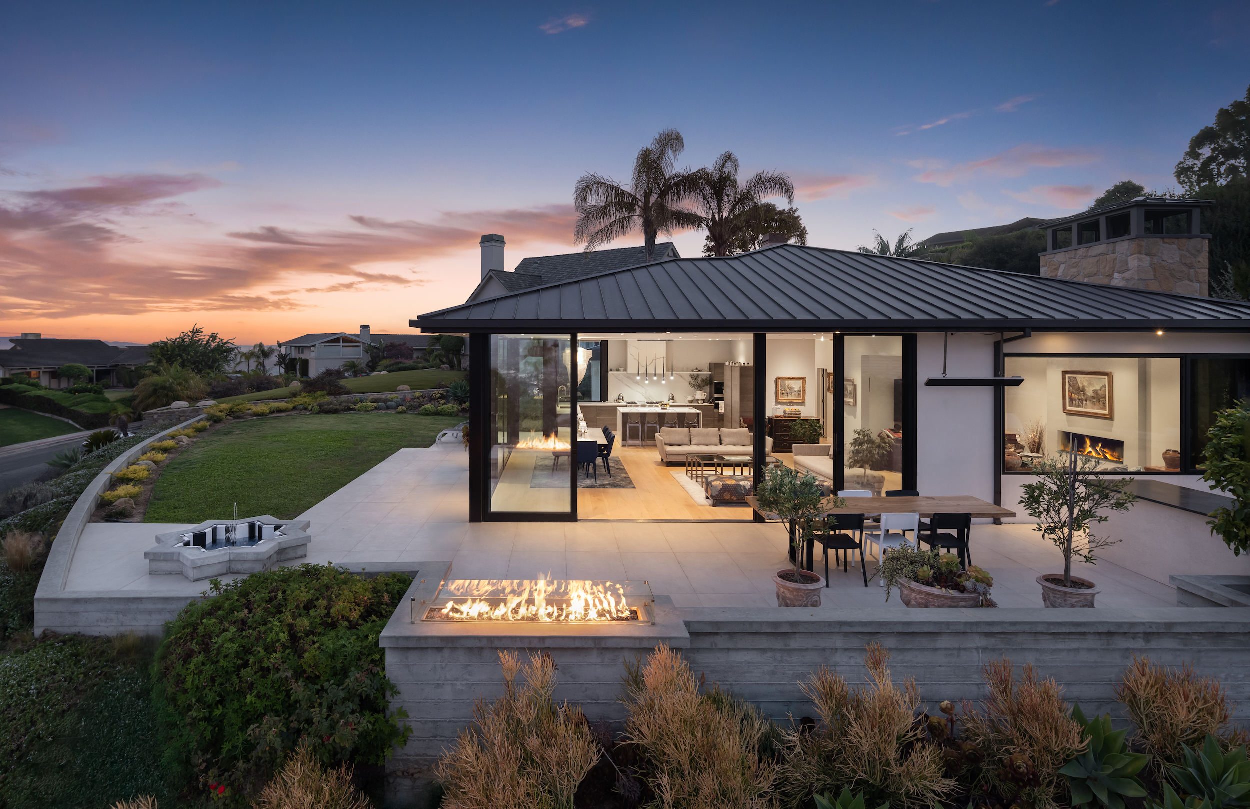 A photo of a house at dusk with the sunset in the background. The house has floor to ceiling windows. One of the windows is open and you can see inside the living room and dining area. In the forefront there is a terrace with an outdoor fire pit that has flames. There is a small water fountain on the left side of the terrace.