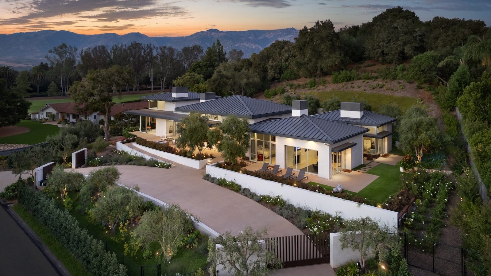 Aerial view of a contemporary hillside residence at sunset, featuring low-profile metal roofs, landscaped gardens, and outdoor terraces framed by mountain views