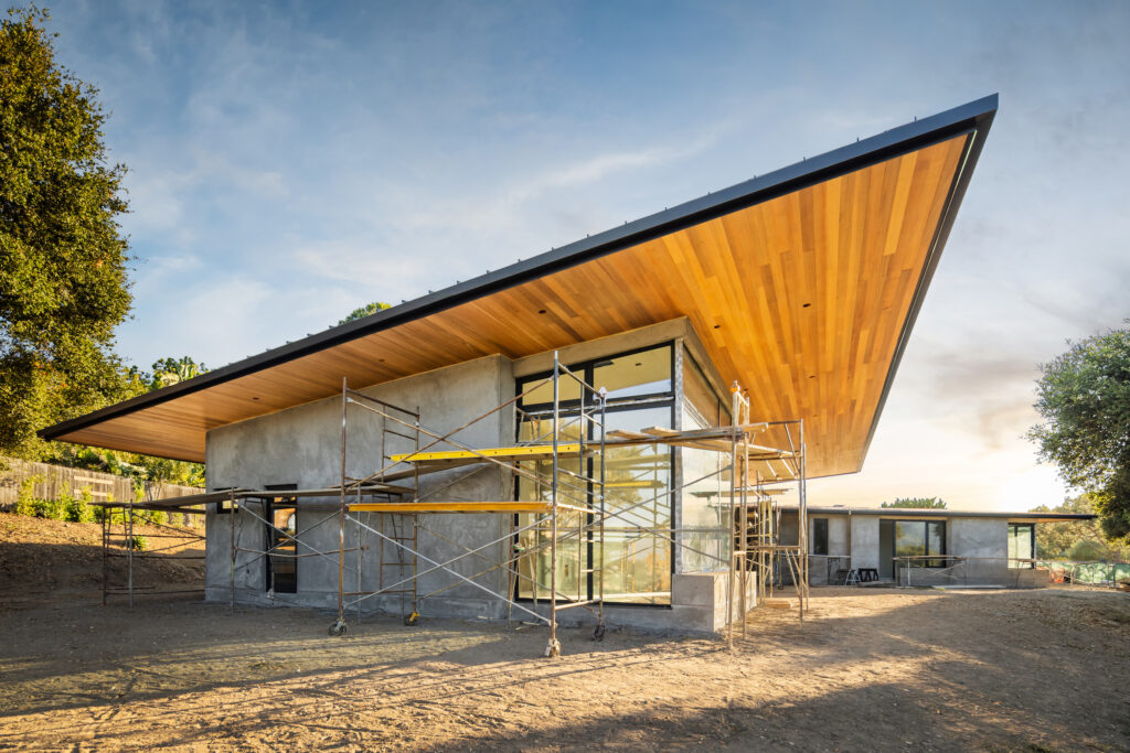 Low-angle shot of a modern house under construction, highlighting a large, sharp-angled roof overhang with warm wood paneling and metal scaffolding against a clear sky.