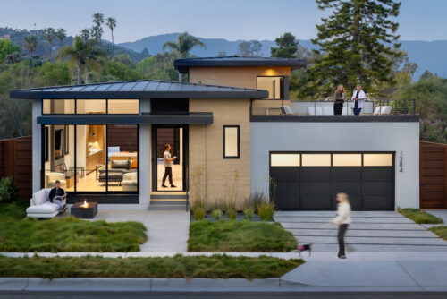 Exterior dusk view of the Shoreline Residence, a modern two-story home with flat roofs and warm wood-paneled accents. The large windows reveal a lit interior, while a person sits by a small fire pit on the front lawn and others stand on the second-story roof deck.