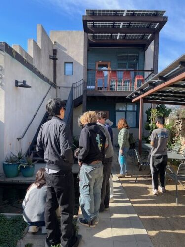 A group of students from Midland School gathers in the sun-drenched outdoor courtyard of Blackbird Architects' Santa Barbara office. They are looking toward a modern, multi-story building featuring a blue-paneled balcony with red chairs, industrial metal stairs, and a shaded outdoor seating area.